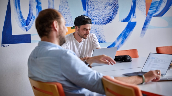 Two system administrators sit in a room with laptops and discuss a project. A wall with a street art motive can be seen in the background.
