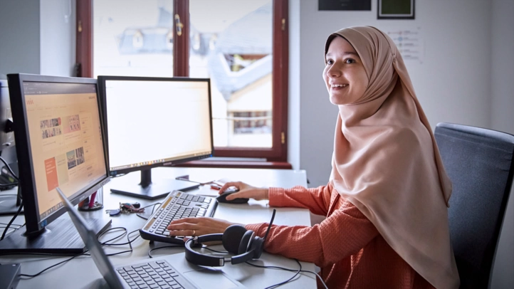 A muslim employee smilingly looks at a person not visible in the picture. She is sitting at a desk with two screens.