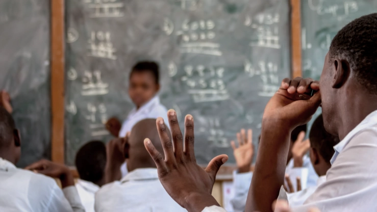 An African school class in the classroom. The teacher, standing in front of a blackboard, looks questioningly into the class. A student raises his hand.