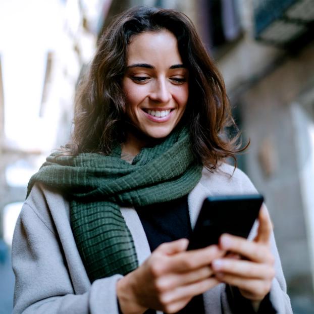 Eine junge Frau mit grauem Mantel und grünem Winterschal in einer schmalen Straße. Sie schaut lächelnd auf ihr Smartphone.