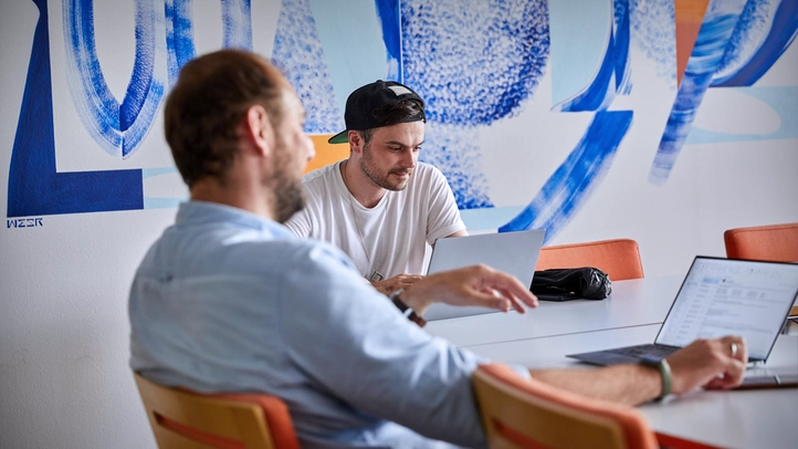 Zwei Mitarbeiter in einem Meetingraum vor Laptops sitzen. Im Hintergrund Streetart an einer Wand.