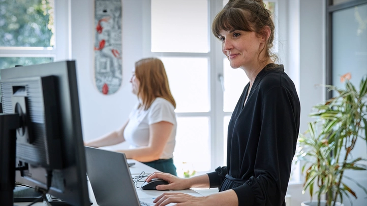 Zwei Mitarbeiterinnen an höhenverstellbaren Schreibtischen arbeitend.