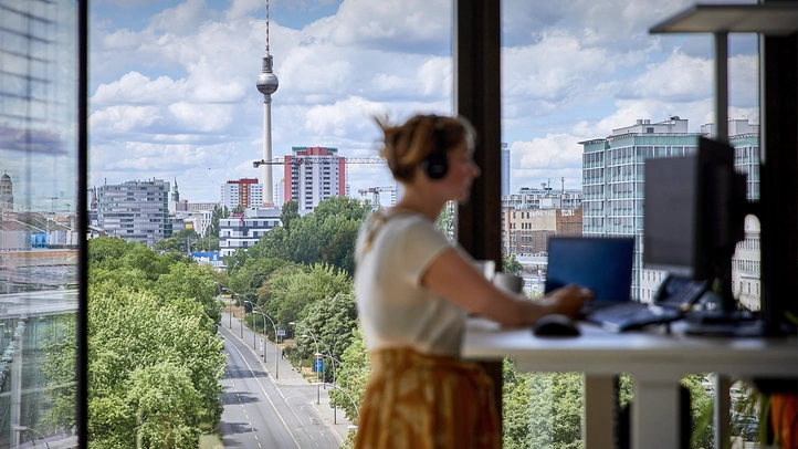 Eine Mitarbeiterin arbeitet stehend am höhenverstellbaren Schreibtisch. Im Hintergrund der Fernsehturm durch ein vollverglaste Fensterfront.