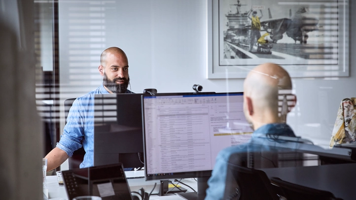 Two employees are sitting across from each other at their workstations and are talking.