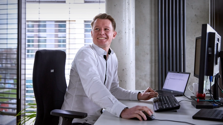 Ein Mitarbeiter mit weißem Hemd erzählt mit einem Kollegen, der nicht im Bild zu sehen ist. Er sitzt im Office am Arbeitsplatz mit PC.