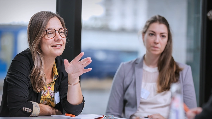 Eine Frau erörtert einen Sachverhalt in einem Workshop des Interop Days. Eine Wissenschaftlerin sitzt zuhörend im Bildhintergrund.