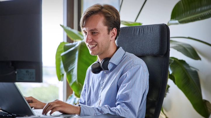 An editor is writing a text on a PC in his office. A large green plant can be seen in the background.