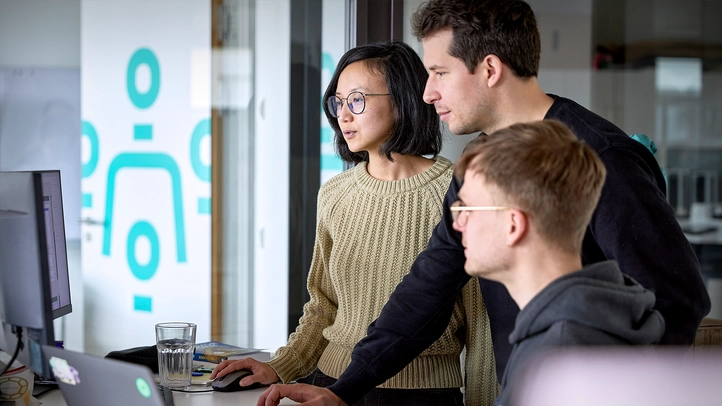 Drei Mitarbeiter:innen besprechen sich am Arbeitsplatz mit Blick auf einen Monitor.