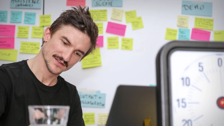 A UX consultant notes down feedback from users in a digitization lab. In the foreground is a timer, in the background a wall with postits.