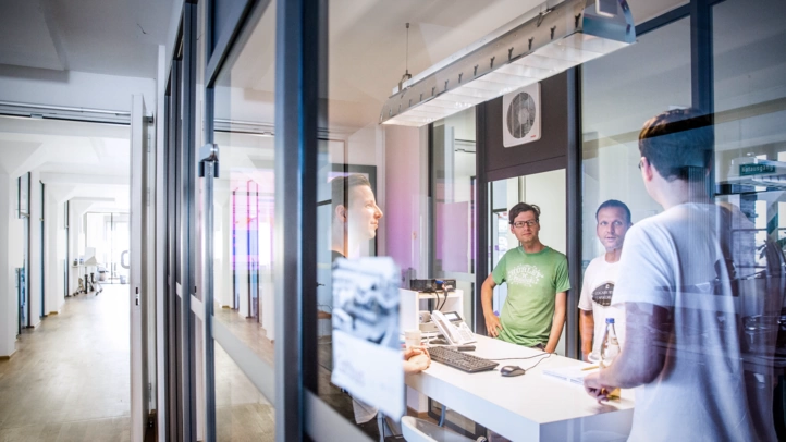 Drei Projektmitarbeiter stehen in einem Besprechungsraum und unterhalten sich. Im Hintergrund sieht man einen Gang mit Büros am Berliner Standort.