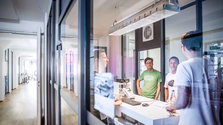 Three project employees are standing in a meeting room and talking. In the background, you can see a corridor with offices at the Berlin site.