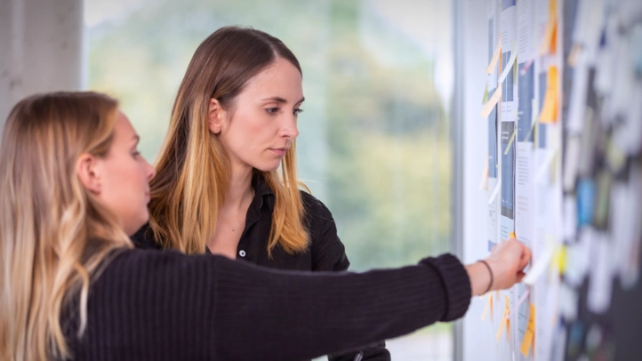 Eine UX-Beraterin und eine Designerin bewerten mithilfe eines Moodboards an der Wand Projektansätze.