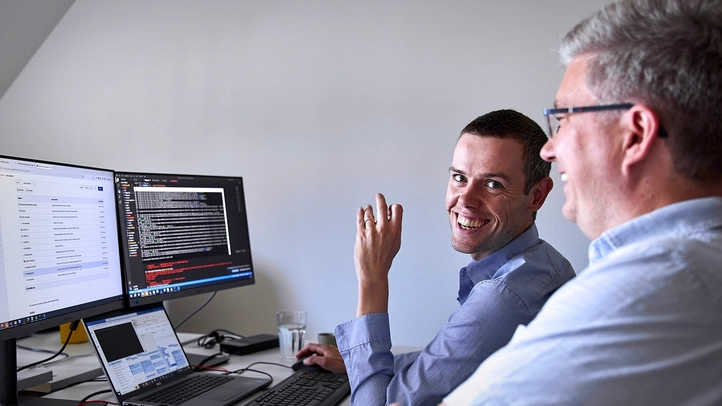 Two colleagues from Leipzig discussing at their workstation. Program code can be seen on the screen.