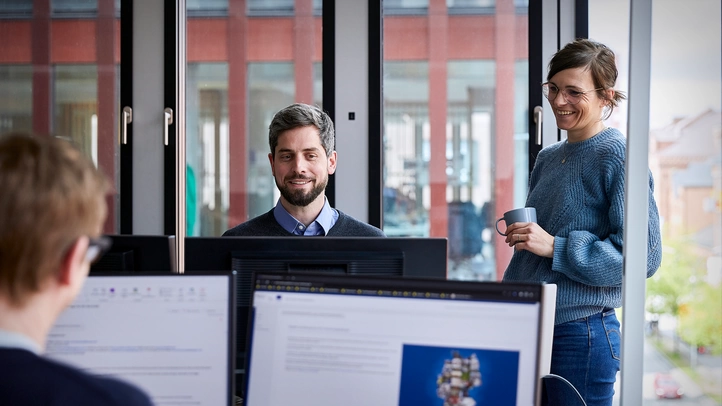 Zwei Mitarbeitende am Hamburger Standort unterhalten sich im Büro. Ein dritter Mitarbeiter arbeitet ihnen gegenübersitzend am Computer.