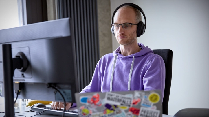 Ein Mitarbeiter mit Kopfhörern arbeitet konzentriert am PC. Sein Notebook ist mit bunten Stickern beklebt.
