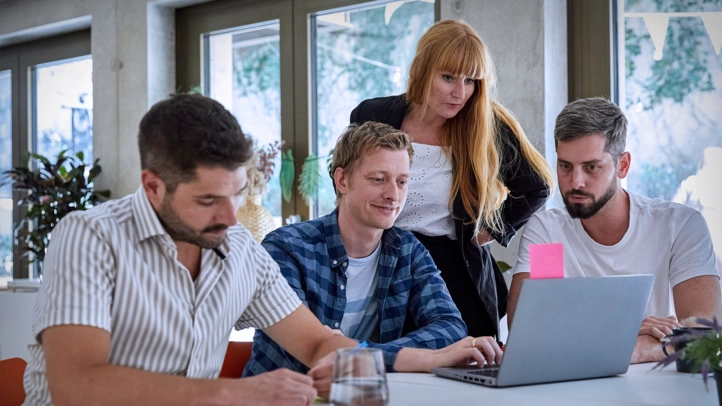 Ein Projektteam bespricht sich in einem großen Besprechungsraum. Alle blicken konzentriert auf einen Laptop und diskutieren miteinander.