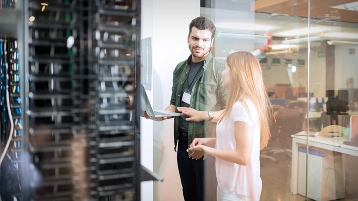 A woman and a man are standing in front of a server cabinet in the data center and are talking. The man is holding an open laptop in front of him.
