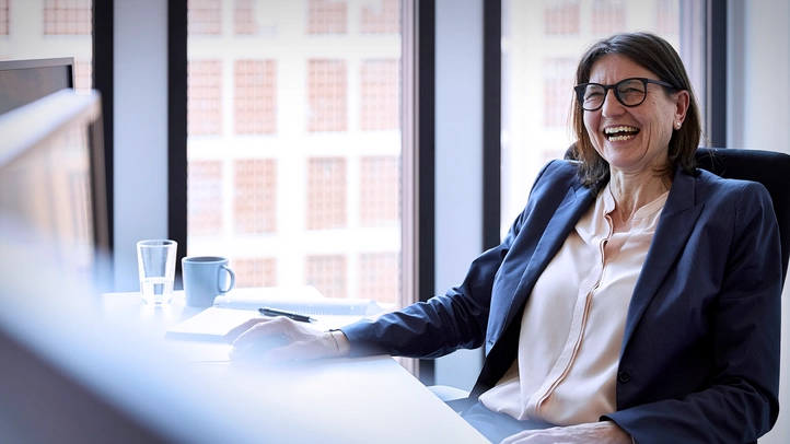 Eine Frau im Business-Kostüm sitzt am Schreibtisch und unterhält sich lächelnd mit einer nicht im Bild sichtbaren Person.