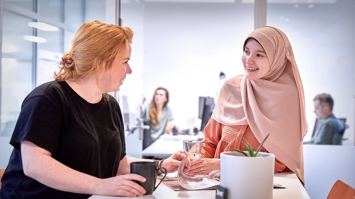 Two female employees are drinking coffee in the atrium at the Mainz site. One person is wearing a headscarf.