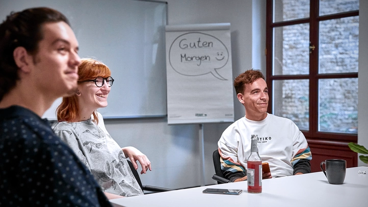 Two employees and one female employee listen to a speaker in a large meeting room at the Mainz site.