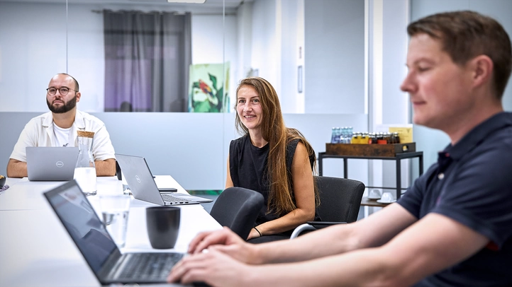 One employee types on his laptop while two employees listen to the presentation of one person.