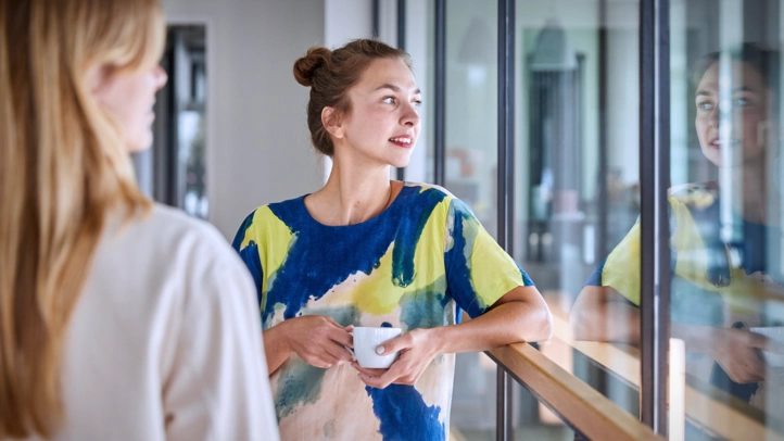 Zwei Mitarbeiterinnen stehen mit Kaffeetassen auf dem Flur und unterhalten sich. Das Gesicht der einen Person spiegelt sich in der Glasfront.