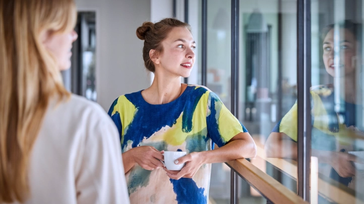 Two employees are standing in the hallway with coffee cups and talking. One person's face is reflected in the glass front.