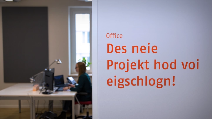 An employee is working at a desk. She was photographed through the door area. Next to the door is the writing "Des neie Projekt hod voi eigeschlogn!" (in Bavarian) can be seen printed in adhesive letters on the wall.