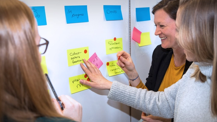 Drei Mitarbeiterinnen aus München vor einem Kanban-Bord. Sie sortieren den Projektstatus.