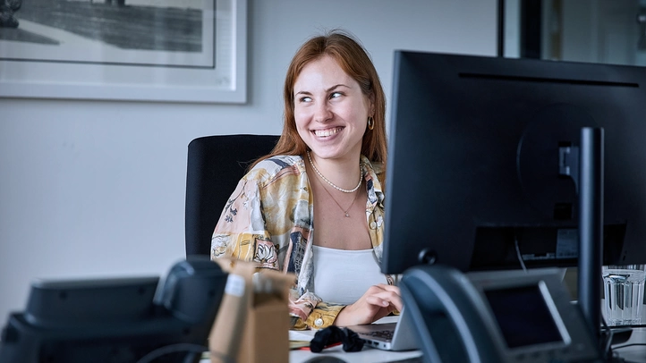 Eine Mitarbeiterin sitzt an ihrem Büroschreibtisch und lächelt einem Kollegen zu, der nicht im Bild zu sehen ist. Im Vordergrund ist ihr Bildschirm zu sehen.