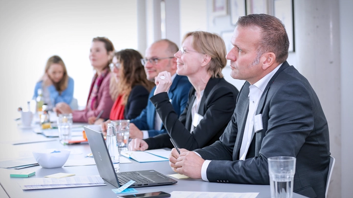 In einem Digitalisierungslabor sitzen mehrere Teilnehmer an einem großen Konferenztisch. Sie hören aufmerksam einer Person zu, die auf dem Bild nicht zu sehen ist.