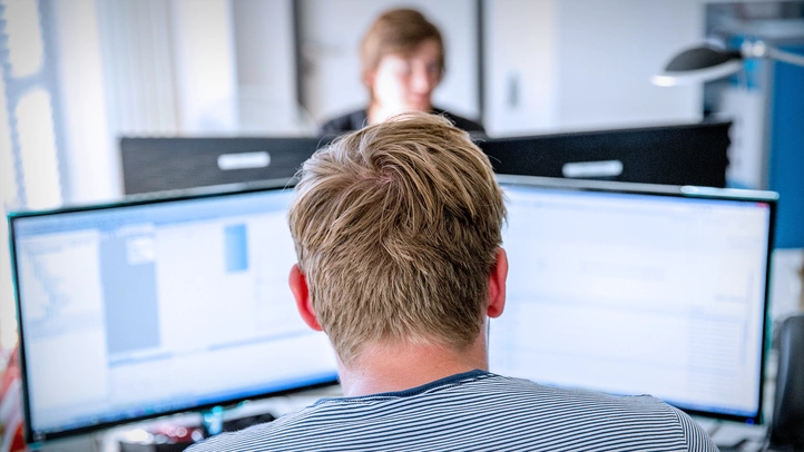 Ein Mann arbeitet am PC-Arbeitsplatz. Im Hintergrund ist eine weitere Person zu sehen.