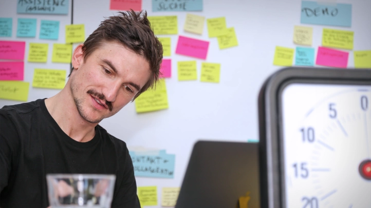 A UX designer writes user stories on Post-its. Next to him is a timer, behind him is a wall covered with colorful post-its.