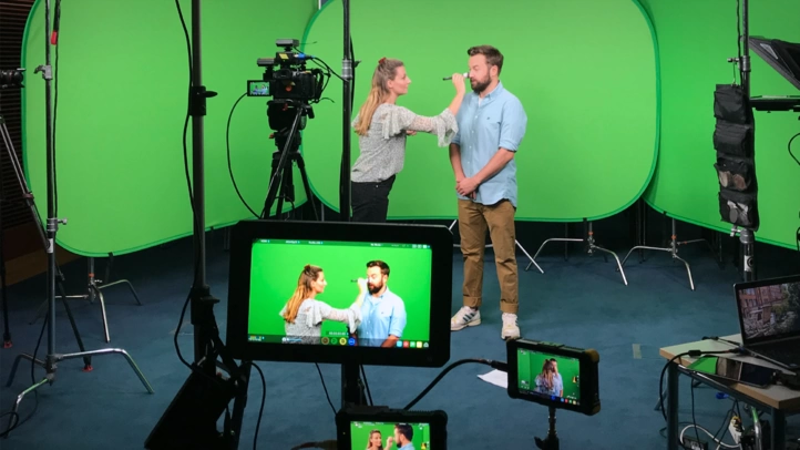 A video producer from ]init[ is powdering an interview partner in front of a green screen. In the foreground, the camera and various monitors can be seen.