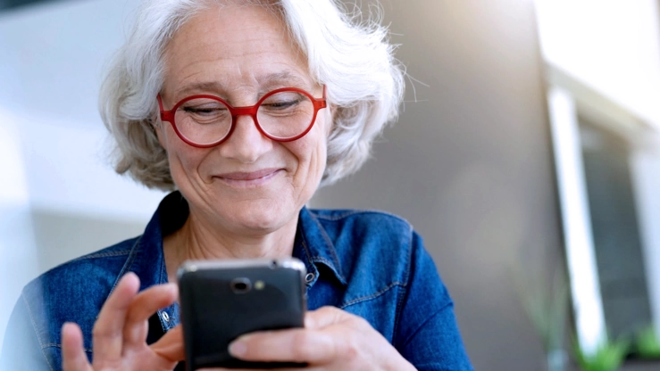 A senior woman with white hair and red glasses scrolls smiling on her smartphone.