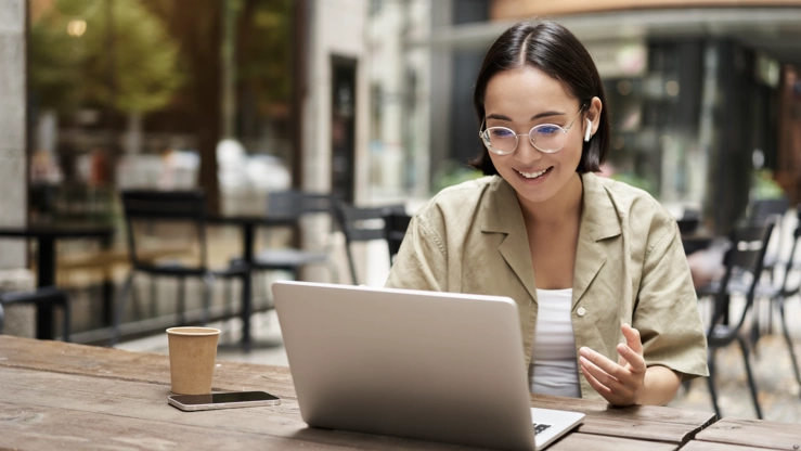 Eine junge Frau sitz im Hof auf einer Holzbank und nimmt an einem virtuellen Meeting am Laptop teil.
