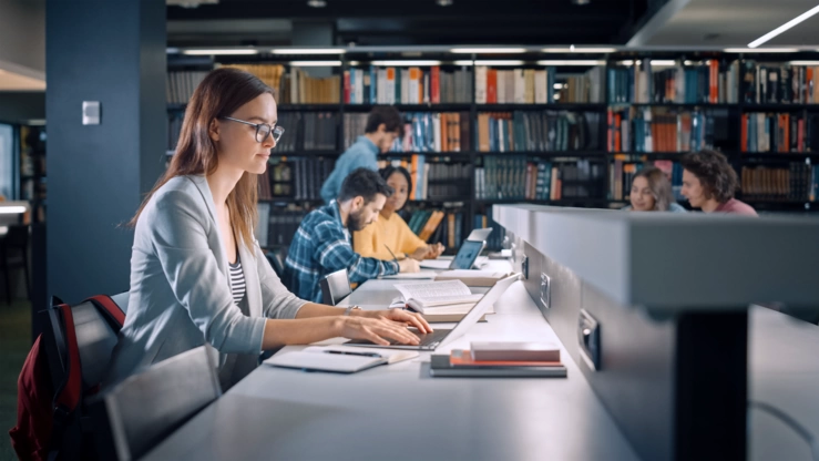 Eine Studentin arbeitet abends in einer Bibliothek am Laptop. Im Hintergrund sind weitere Student:innen vor einem Bücherregal zu sehen.