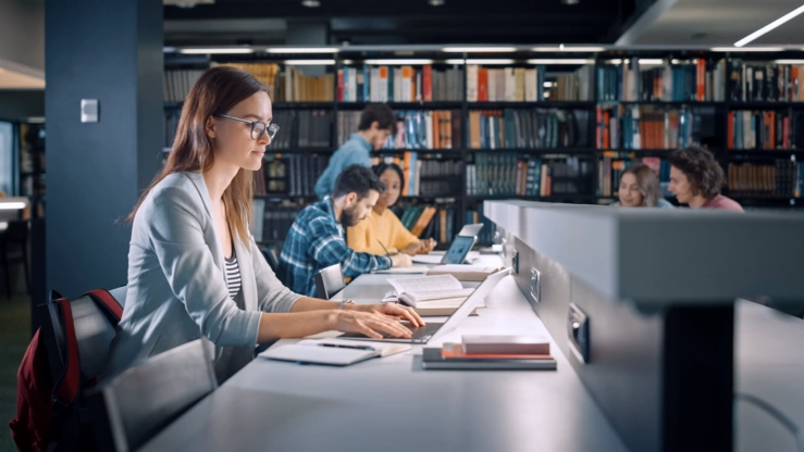 A student is working on her laptop in a library in the evening. In the background, other students can be seen in front of a bookshelf.