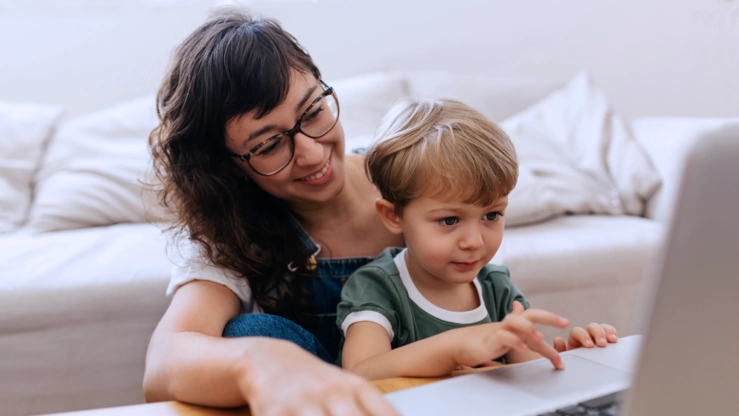 Eine junge Mutter mit ihrem Kind vor einem Laptop. Der Junge navigiert auf dem Touchpad.