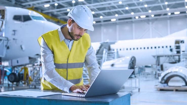 An aviation expert wearing a high-visibility vest and helmet works on his laptop in a hangar. In the background, aircraft can be seen being assembled and maintained.