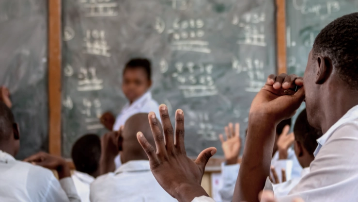 Eine afrikanische Schulklasse im Unterricht. Die Lehrerin, vor einer Tafel stehend, schaut fragend in die Klasse. Ein Schüler meldet sich.