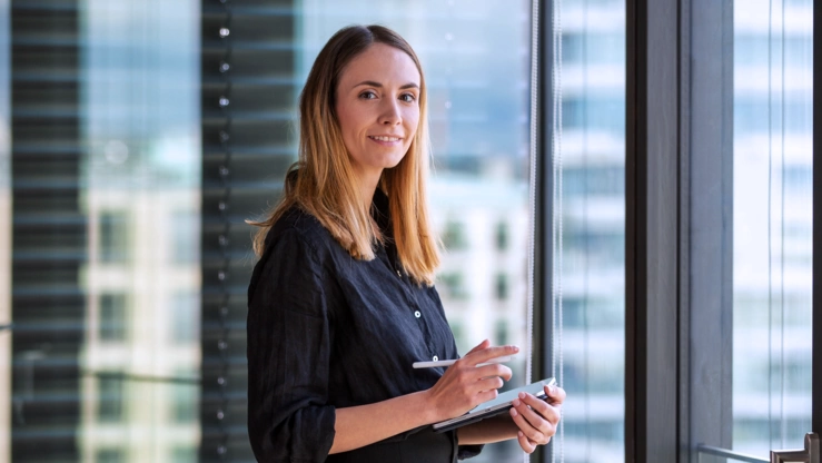 Die UX Designerin Nicole Lössner steht mit einem Tablet im Office. Im Hintergrund ist die Innenstadt Berlins zu sehen.