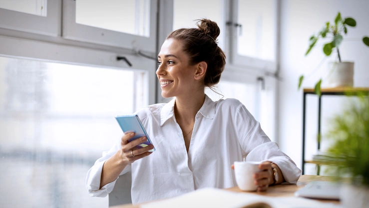 Frau mit Smartphone schaut inspiriert aus dem Fenster