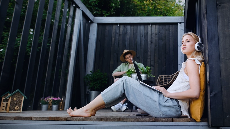 A young woman sits on a porch with a laptop and headphones. In the background, a teenager tends green plants.