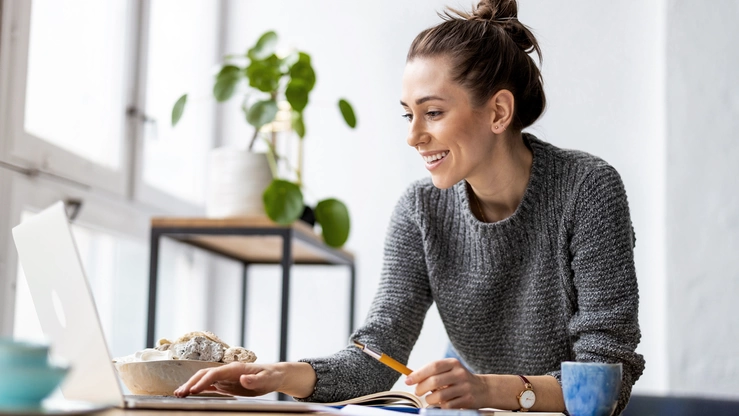 A young woman looks at her laptop. She is sitting in a light-filled room with a cup of coffee next to her on the desk.