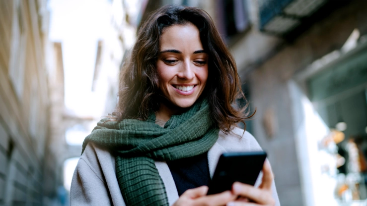 A young woman with a gray coat and a green winter scarf in a narrow street. She looks at her smartphone with a smile.