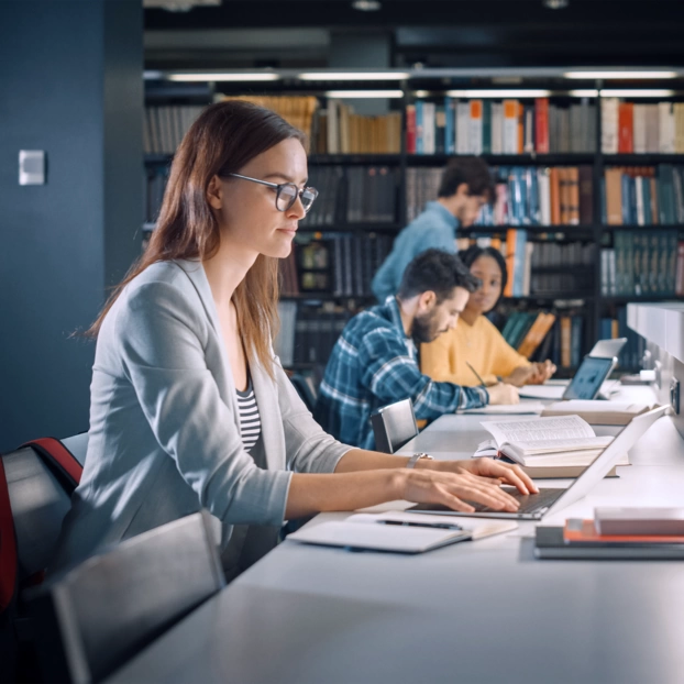 A student is working on her laptop in a library in the evening. In the background, other students can be seen in front of a bookshelf.