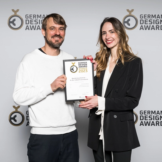 Nicole Lössner und Philipp Hahn von ]init[ mit der Urkunde des German Design Award vor einer Fotowand bei der Preisverleihung.