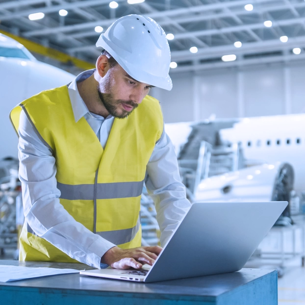 An aviation expert wearing a high-visibility vest and helmet works on his laptop in a hangar. In the background, aircraft can be seen being assembled and maintained.