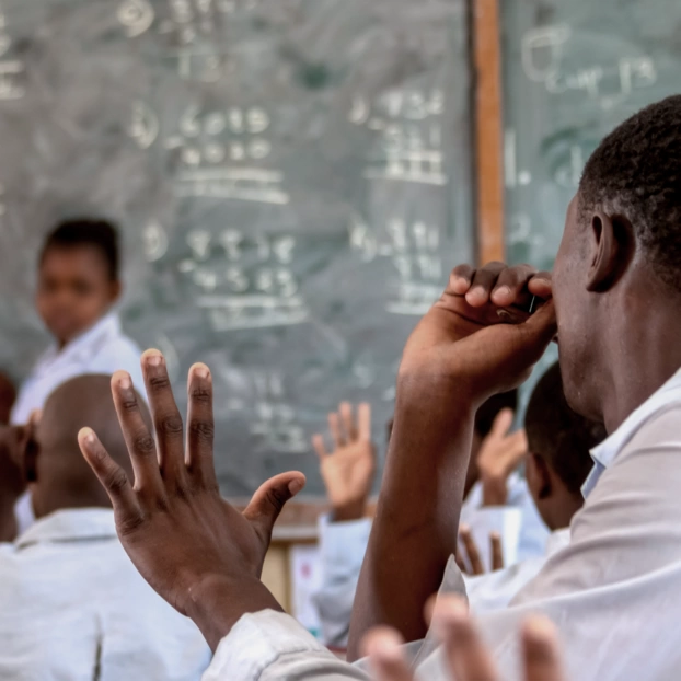An African school class in the classroom. The teacher, standing in front of a blackboard, looks questioningly into the class. A student raises his hand.